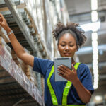 Operadora de logística realizando picking em estoque de armazém usando tablet para separar pedidos nas prateleiras
